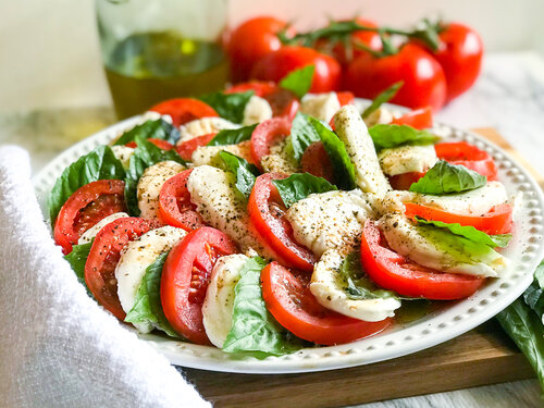 Caprese Salad | Keto, Low-Carb - Photo of a Caprese Salad arranged on a white plate atop a wooden cutting board - ready to serve. Additional tomatoes on the vine and extra virgin olive oil appear in the background. Photographed by Adrianna Ruff for AGirlCalledAdri