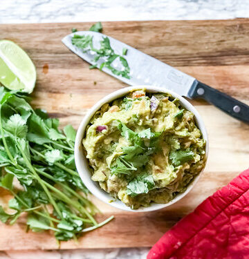 Guacamole in a white bowl next to cilantro and lime on a cutting board