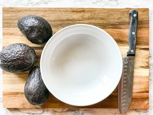 three uncut avocados next to a white bowl and knife on a wooden cutting board