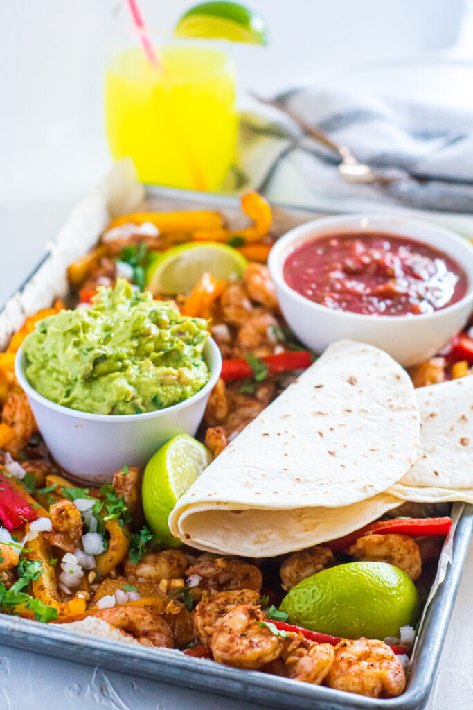 sheet pan blackened shrimp fajitas photographed with bowls of salsa and guacamole with drink in background.