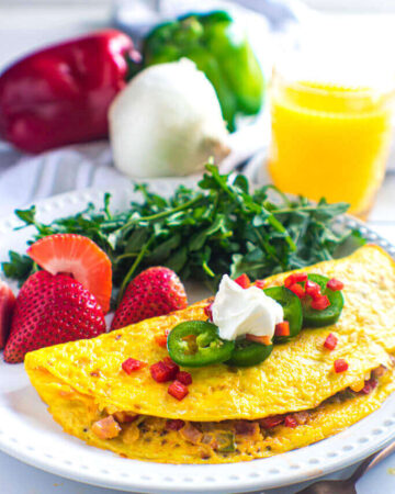 Denver omelet plated with strawberries and arugula. Orange juice and bell peppers are in the background. Omelet is topped with jalapeno, diced red bell pepper, and sour cream.