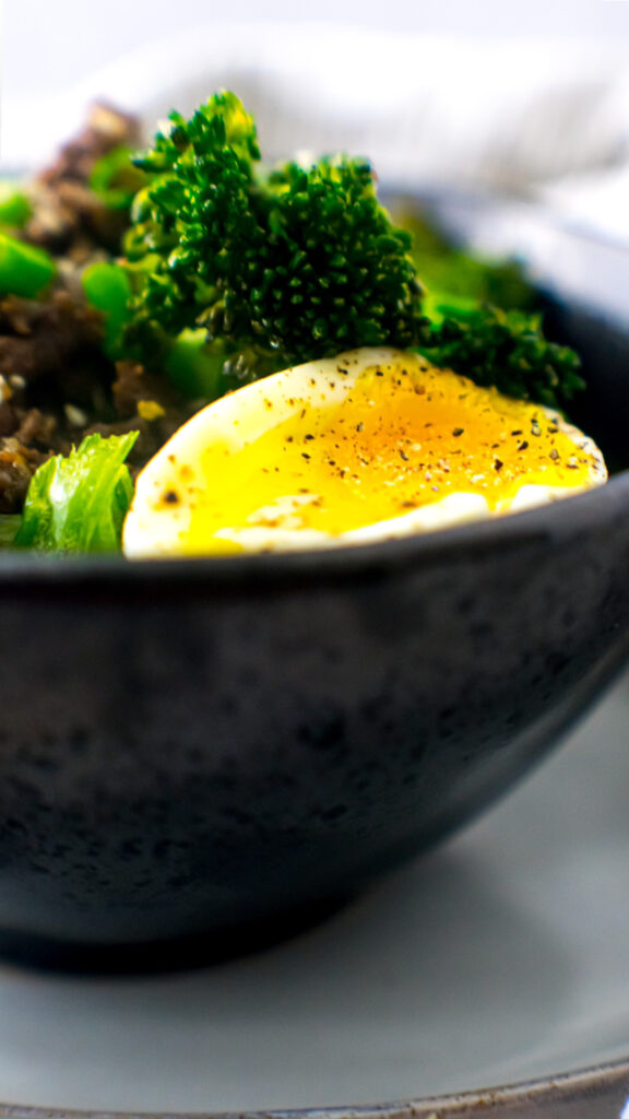 Ground beef bulgogi in a black bowl with broccolini, soft boiled egg, green onions, and toasted sesame seeds.