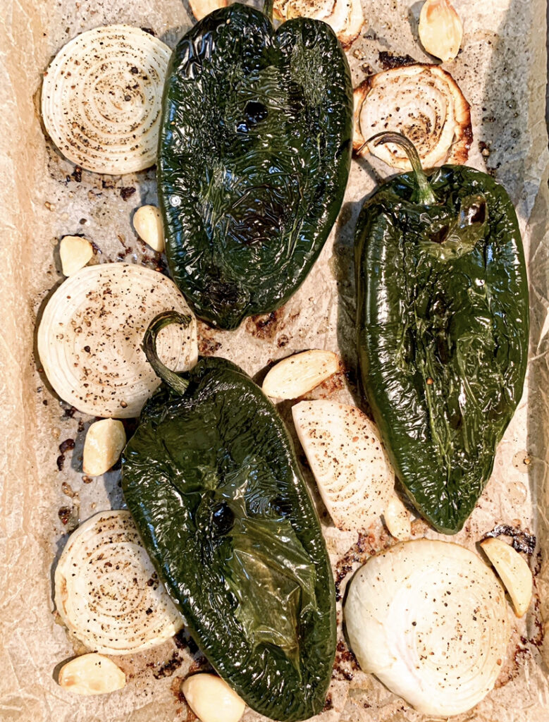 Roasted green poblano peppers, sliced yellow onions, and peeled garlic on a parchment paper-lined baking sheet.